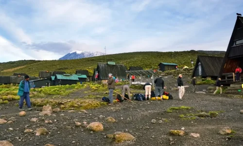 Hikers-are-preparing-gear-in-a-rocky-clearing-surrounded-by-dark-A-frame-huts-at-Horombo-Hut-with-the-snow-capped-peak-of-Kilimanjaro-visible-in-the-back